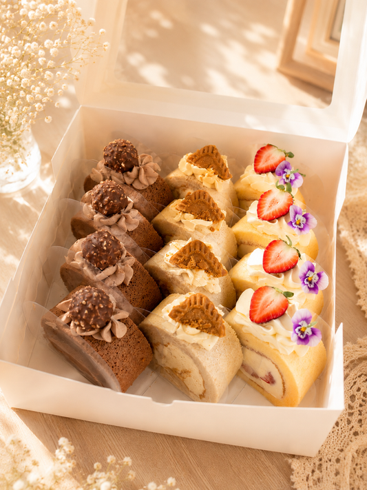 A white bakery box filled with assorted pastries, including chocolate rolls, cream-filled cakes, and strawberry-topped slices, styled on a beige surface with soft decorative flowers in the background.