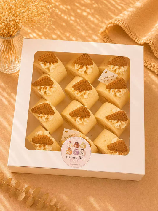 A white square box containing 12 mini Cloud Roll cakes, arranged in a grid with a clear window. Each cake is topped with a small triangular biscuit and a swirl of cream.