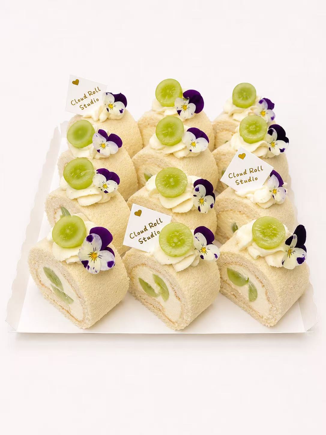 Six cucumber and white chocolate swiss roll cakes arranged on a white rectangular tray against a clean white background. Each cake is topped with a slice of green cucumber, a white chocolate flower, and a purple pansy, with a small paper flag reading 'Cloud Bell Studio'.