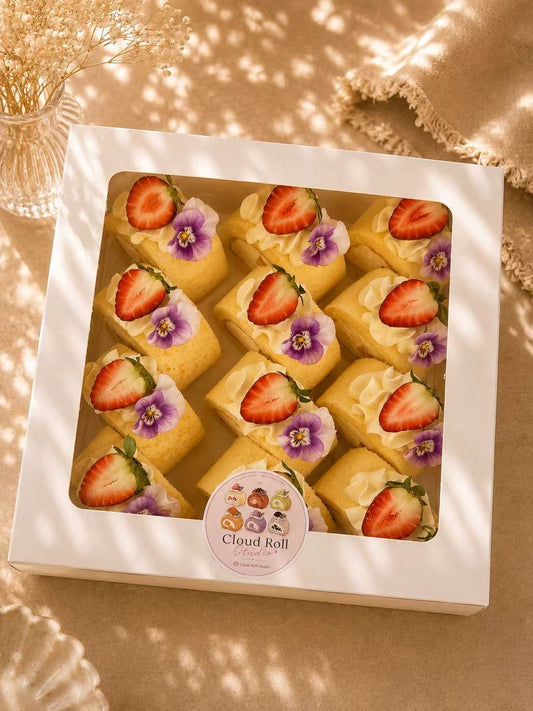 A white square gift box containing several yellow cloud rolls topped with sliced strawberries and purple edible flowers, photographed on a warm beige surface with soft dappled shadows. A round sticker on the front reads 'Cloud Roll'.