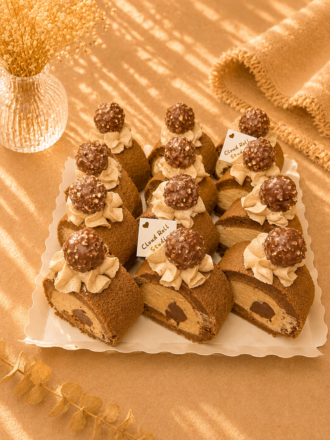 A tray of chocolate swiss roll pastries topped with whipped cream and chocolate spheres, arranged on a white scalloped platter against a warm beige fabric background with decorative dried flowers.