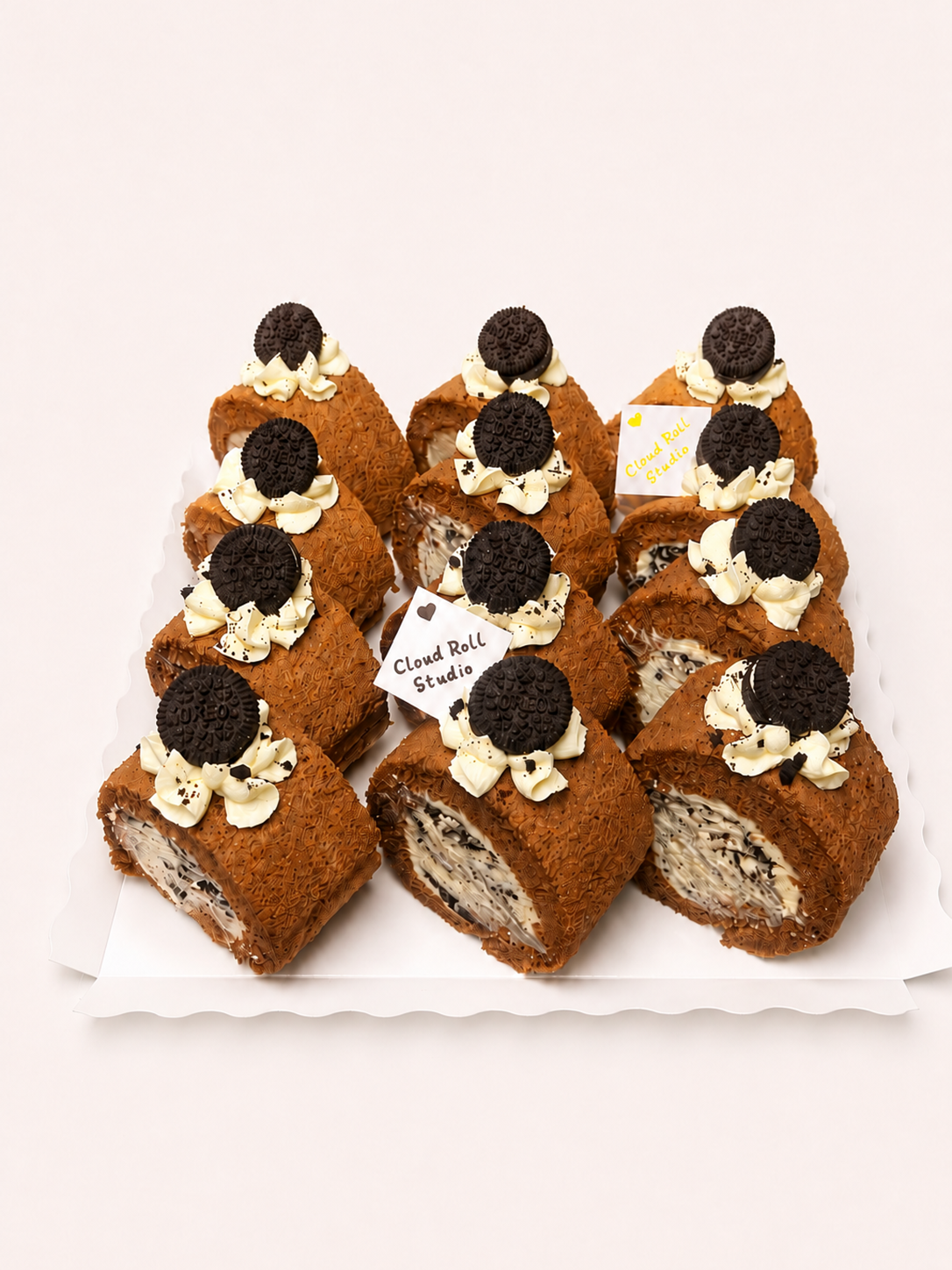 A set of chocolate cake rolls decorated with cream and dark cookie toppings, arranged on a white scalloped tray against a light beige background. One tag reads 'Cloud Butt Studio'.
