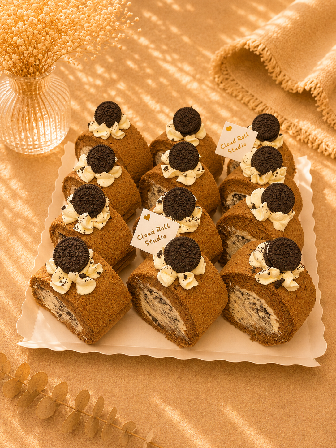 A tray of chocolate cake rolls with white cream and dark cookie toppings, arranged on a white plate with decorative dried flowers and fabric in a warm golden setting.