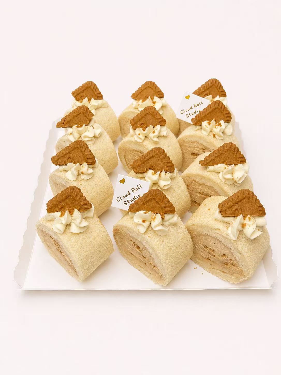 A set of ten cream-filled pastry rolls decorated with brown cookie pieces on top, arranged on a white rectangular plate against a plain white background.