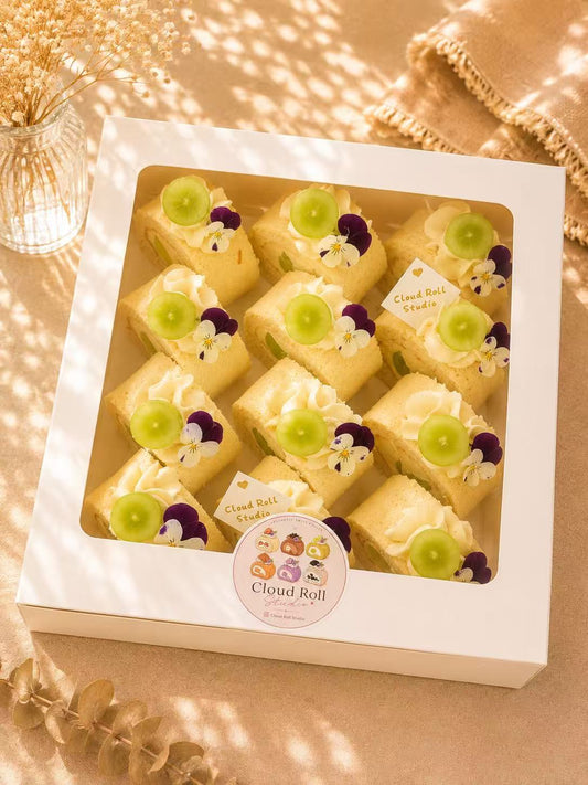 A white square box containing six decorated Cloud Roll cake slices, arranged in a window display. The cakes are topped with whipped cream, green grapes, and edible flowers.