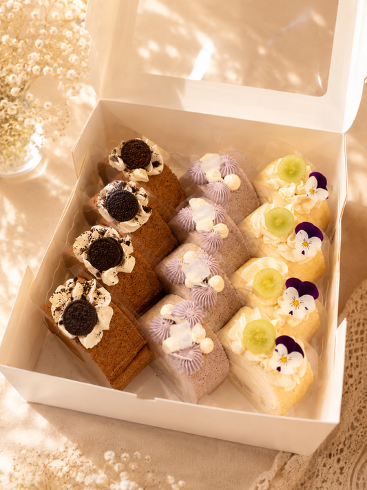 An open white bakery box containing six assorted decorated cake slices or pastry pieces, arranged neatly in a warm, sunlit setting with a soft blurred floral background.