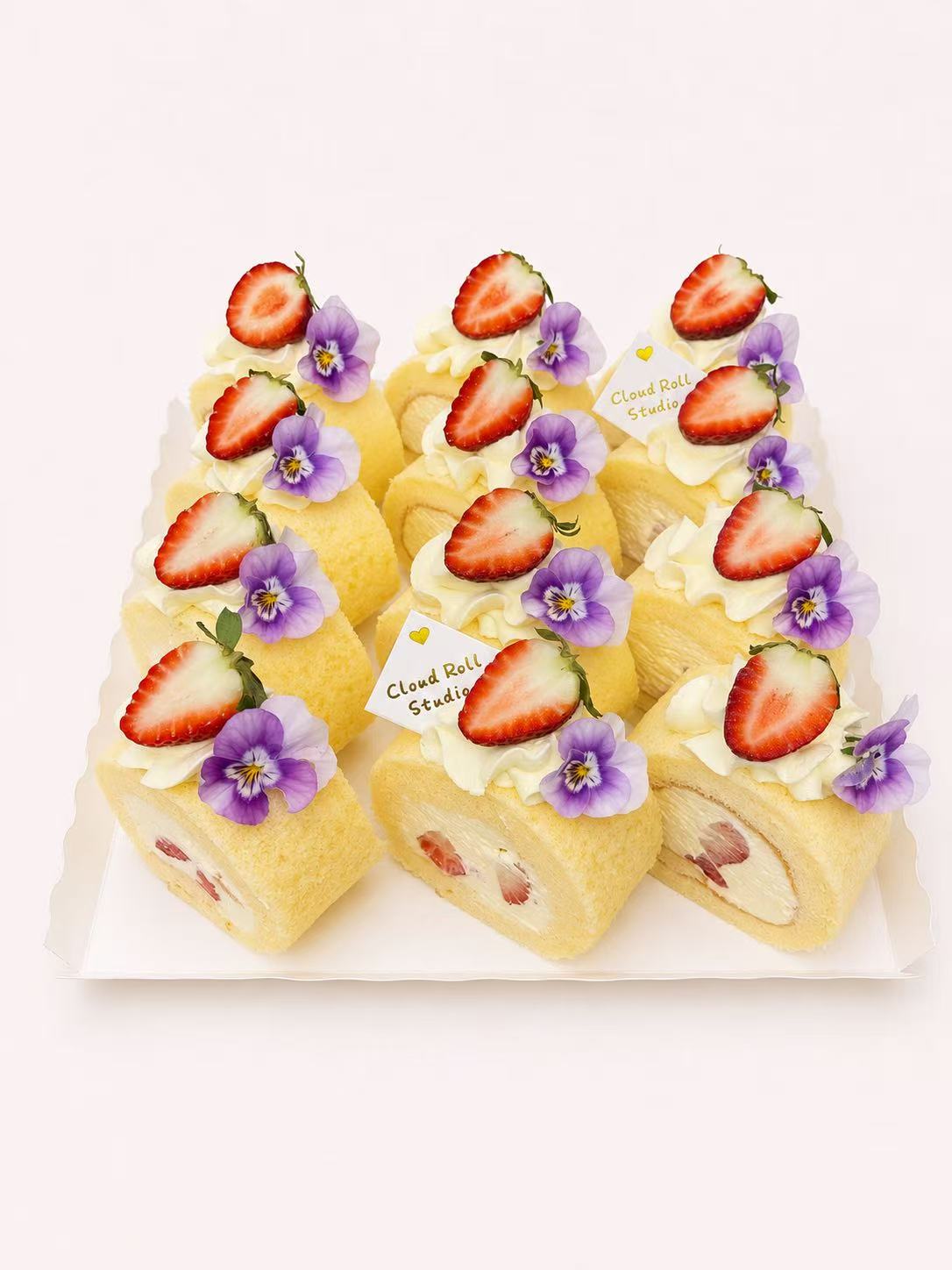 Six strawberry cake rolls arranged on a white scalloped serving tray against a clean white background. Each roll is topped with whipped cream, sliced strawberries, and purple edible flowers, with small tags attached.