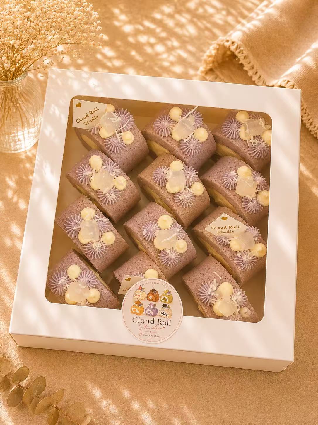 A white square gift box containing multiple Cloud Roll cake slices, photographed from above on a warm beige surface with soft shadows and decorative dried flowers. The box has a clear window and a round Cloud Roll sticker label on the front.