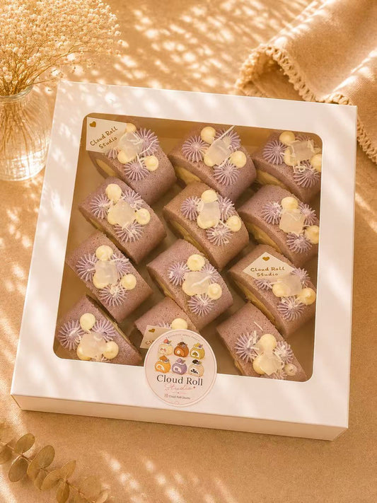 A white square gift box containing multiple Cloud Roll cake slices, photographed from above on a warm beige surface with soft shadows and decorative dried flowers. The box has a clear window and a round Cloud Roll sticker label on the front.