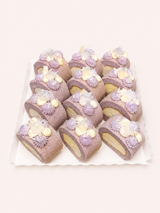 A set of ten small cake rolls decorated with purple frosting swirls, white cream, and translucent flower-shaped candies, arranged on a white scalloped tray against a pale pink background.